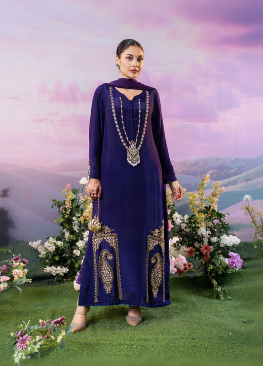 A woman models a deep purple long kurta with gold embroidery and a matching dupatta, standing on artificial green grass with floral arrangements and a scenic backdrop.