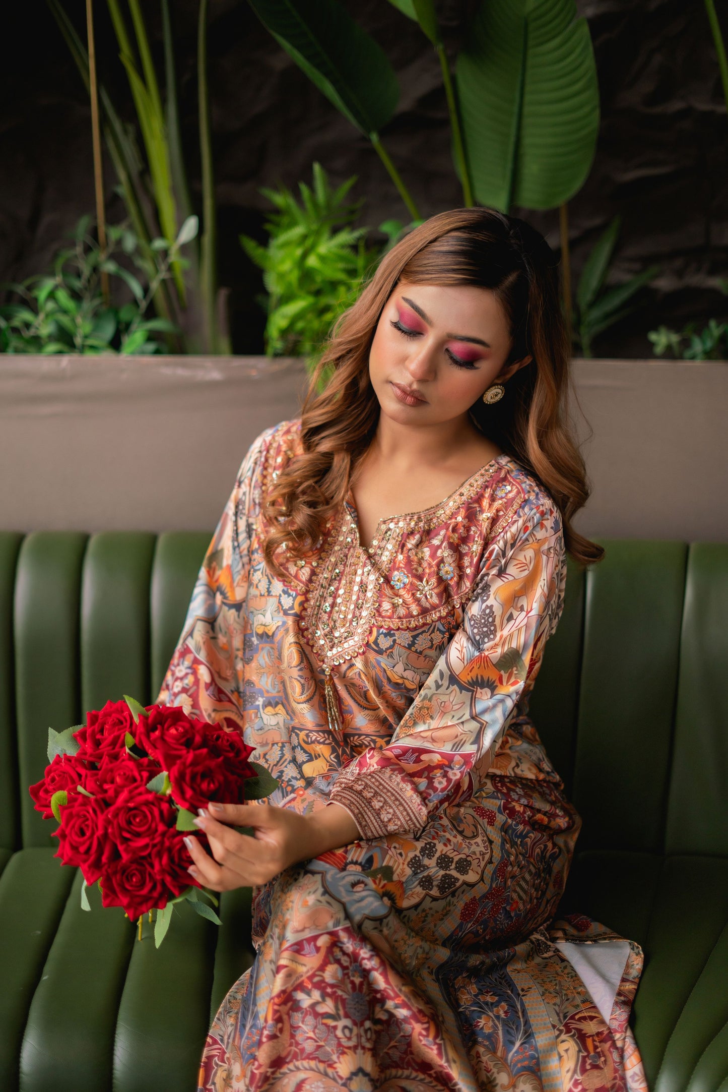Woman in a patterned dress holding red flowers, sitting on a green couch with a blurred natural background